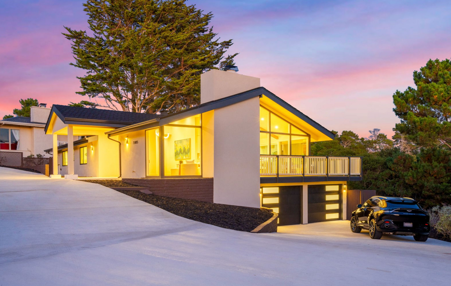 Modern house with large windows lit from within, featuring a two-car garage and a sloped driveway, showcases a Full Interior-Exterior Home Remodel in Carmel, with a car parked outside at sunset.