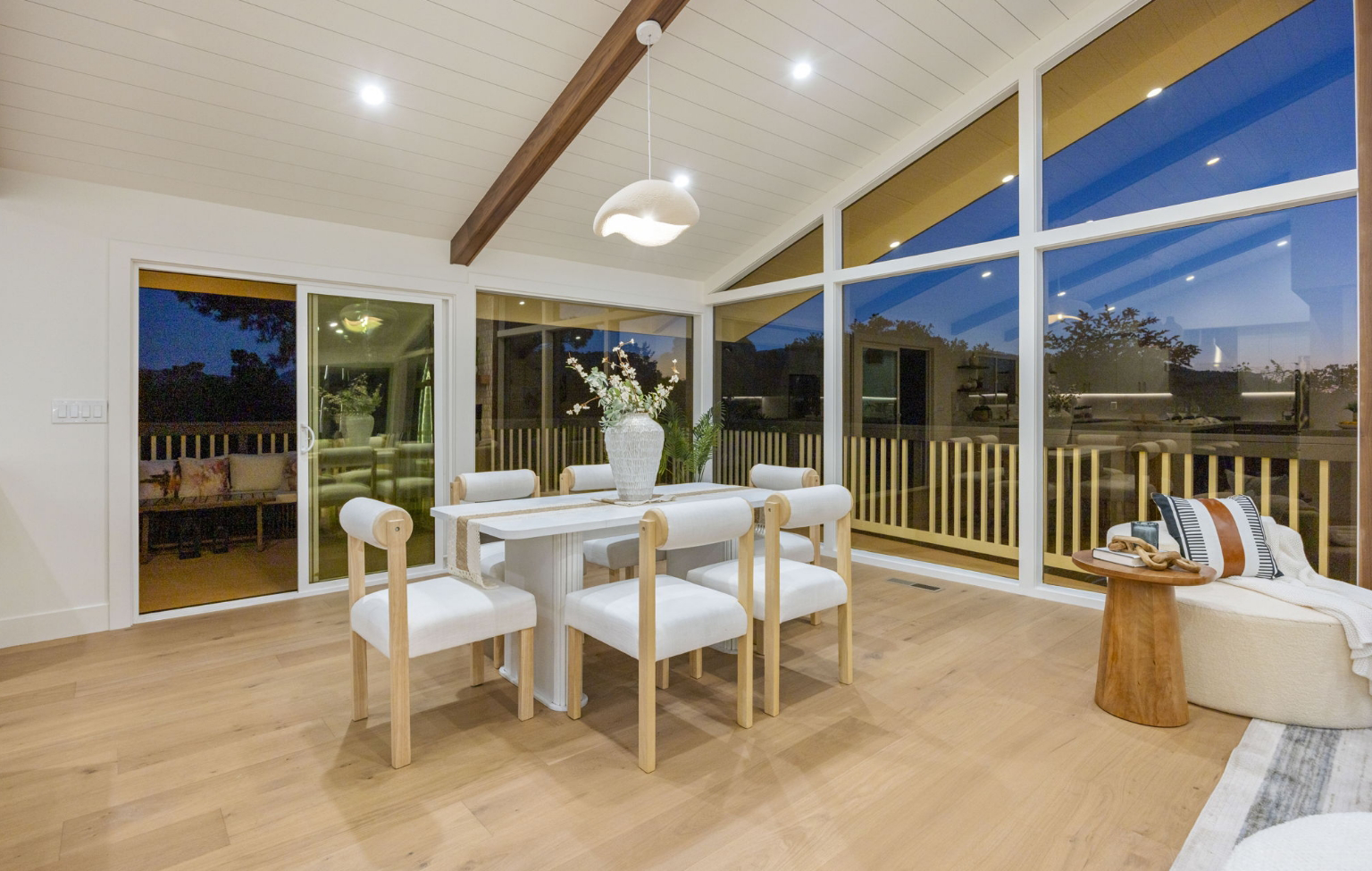 Modern dining area with a white table, six cushioned chairs, large windows, vaulted ceiling with exposed beam, and wood flooring. Part of a Full Interior-Exterior Home Remodel in Carmel; outdoor deck visible through glass.