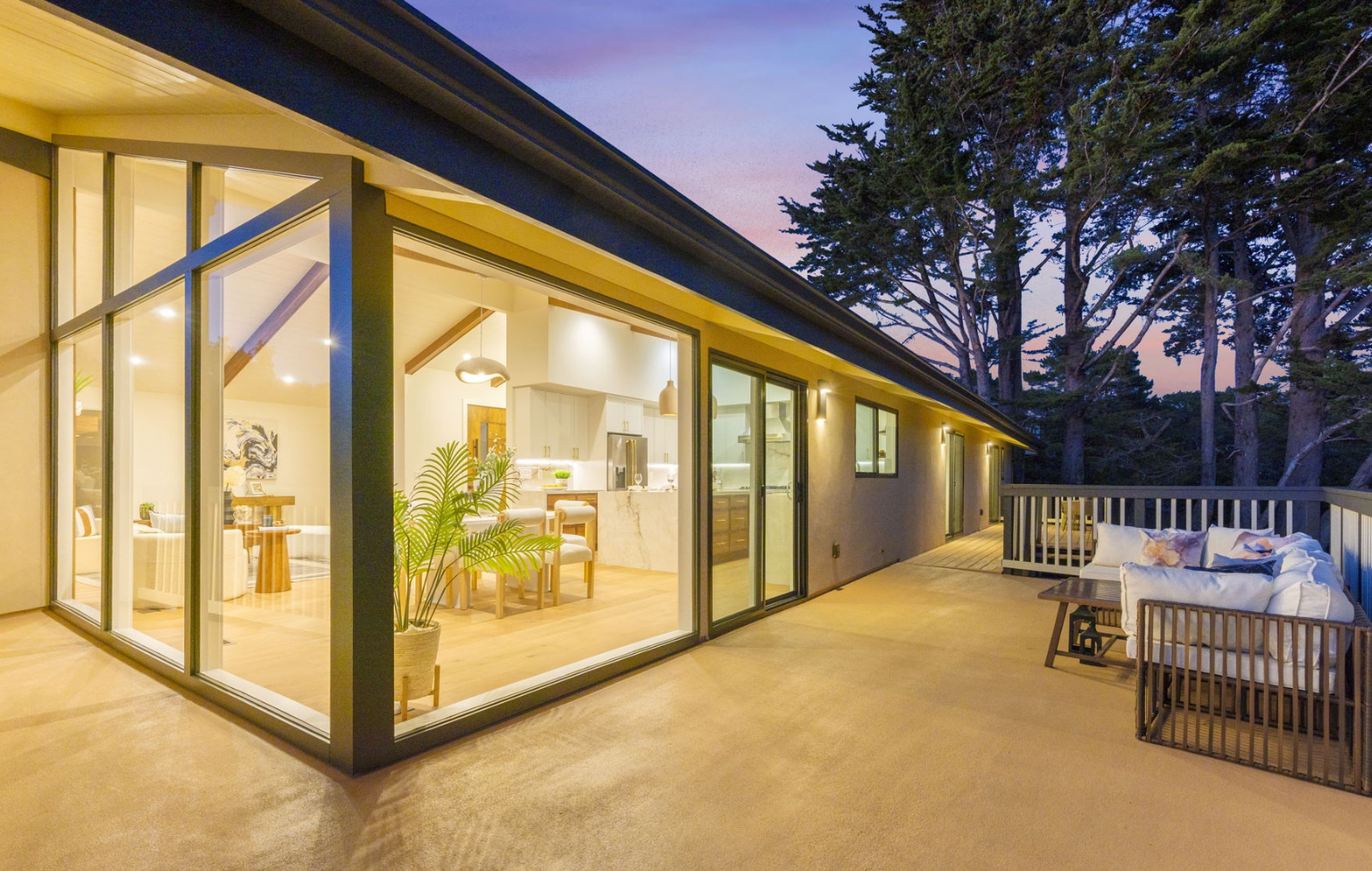 Modern house with large glass windows, open dining area, and a spacious patio with outdoor seating—this Full Interior-Exterior Home Remodel in Carmel is surrounded by tall trees at sunset.