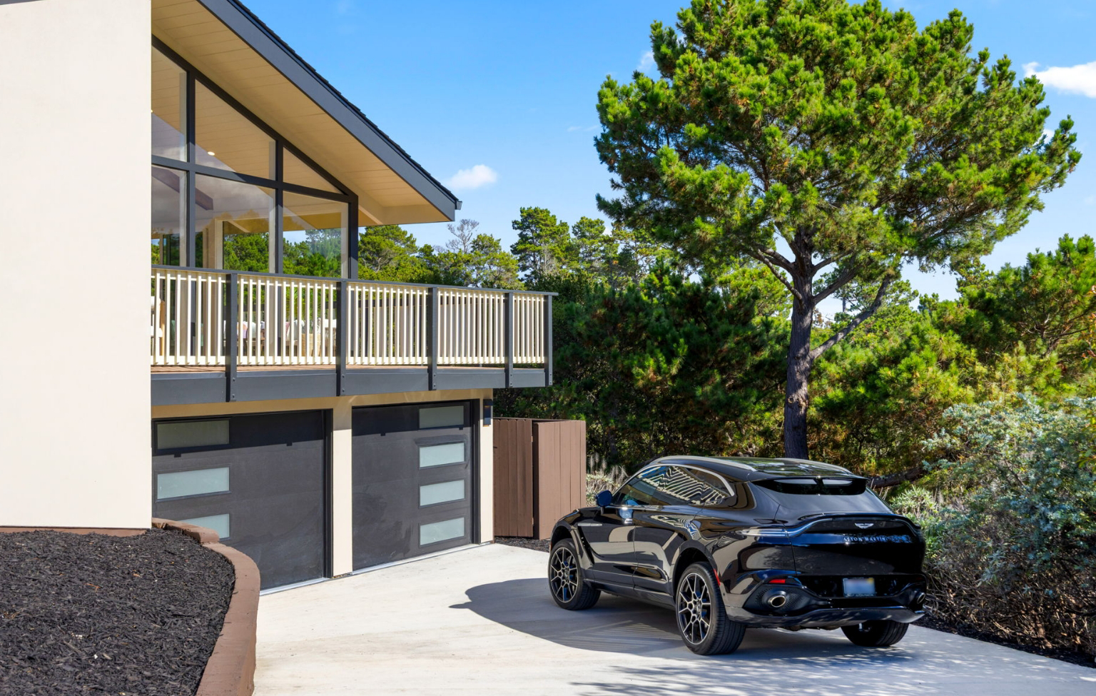 A black SUV is parked in the driveway of a modern house with large windows and a balcony—showcasing a Full Interior-Exterior Home Remodel in Carmel, nestled among trees and clear skies.