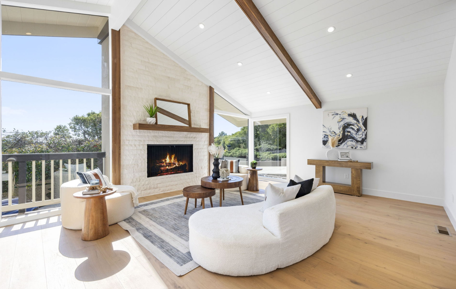 Modern living room with vaulted ceiling, light wood floors, white walls, a stone fireplace, cozy seating, and large windows overlooking greenery—all part of a Full Interior-Exterior Home Remodel in Carmel.