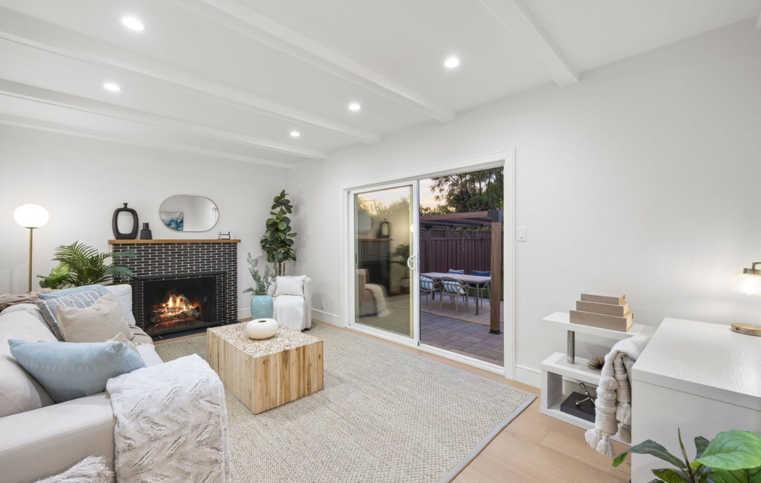 Modern living room with a lit fireplace, light-colored furniture, and sliding glass doors leading to an outdoor patio at sunset.