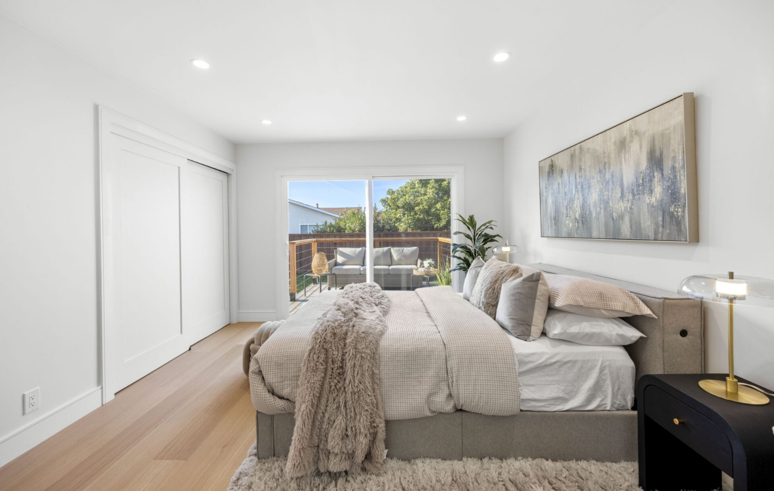 Modern bedroom with neutral bedding, a large painting above the bed, sliding glass doors leading to a patio, light wood floor, and minimal decor.