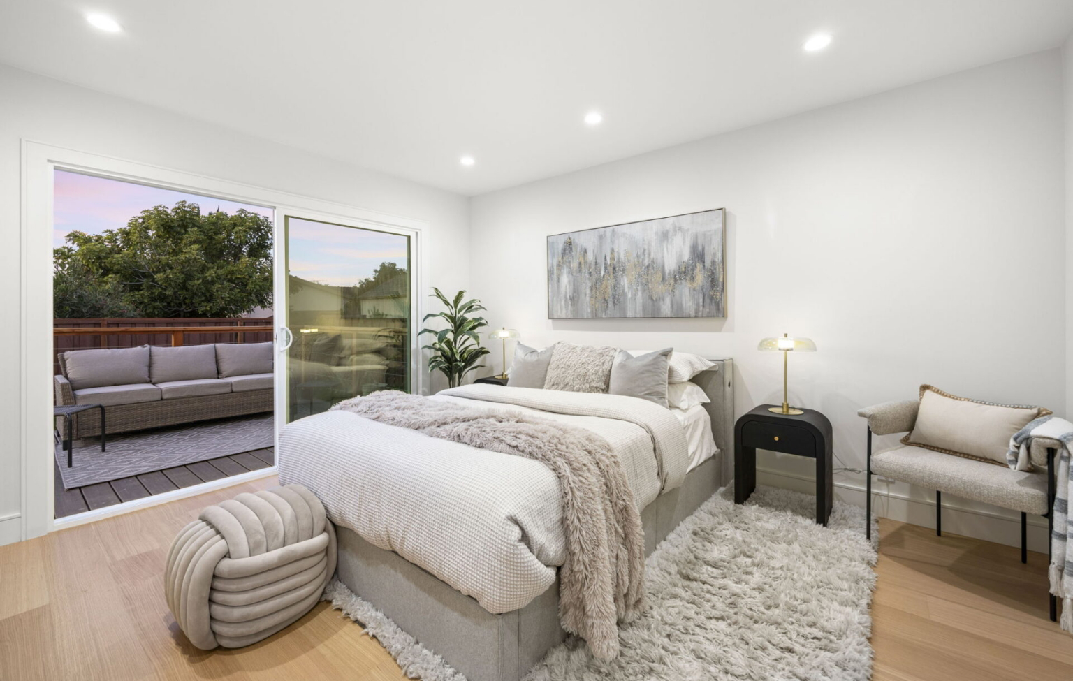 Modern bedroom with a large bed, soft neutral bedding, and a cozy rug, featuring sliding glass doors leading to an outdoor patio with seating and greenery visible outside.