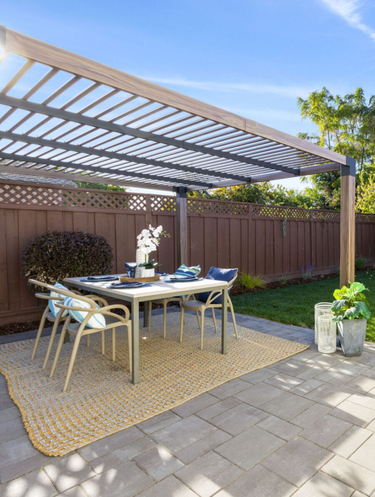 A modern outdoor patio with a slatted pergola, dining table set for four, chairs, decorative plants, and a yellow rug on a paved surface.