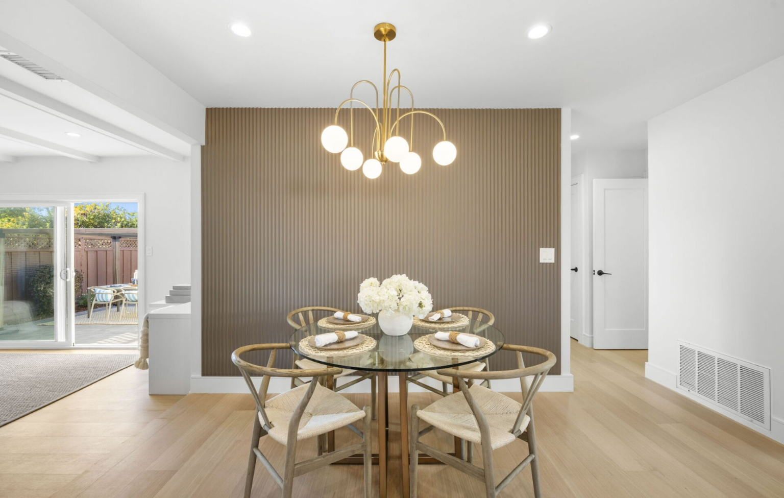 A modern dining area with a round glass table, four wooden chairs, a floral centerpiece, and a contemporary chandelier against a textured accent wall. Sliding doors lead to an outdoor space.
