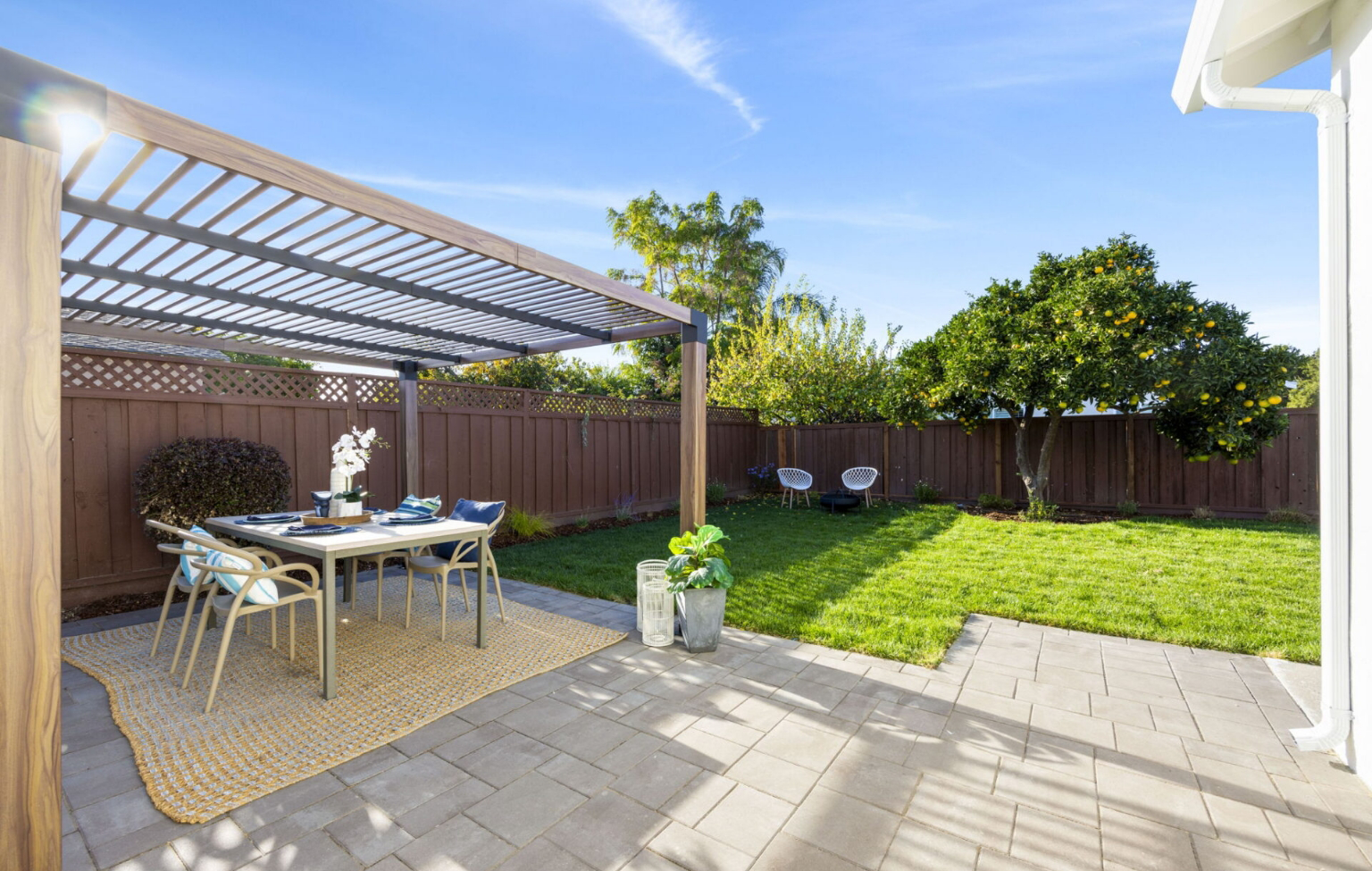 A backyard with a pergola-covered dining area, a grassy lawn, two white chairs under a tree, and a wooden fence under a clear blue sky.