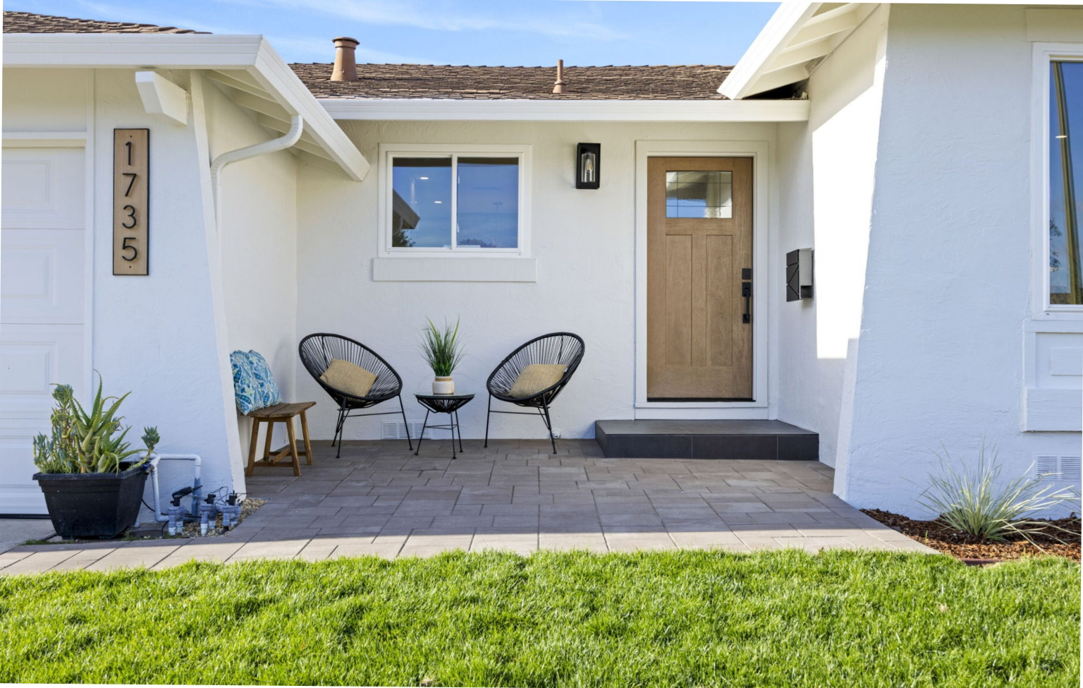 Modern house front entrance with a wooden door, two black chairs with cushions, a small table, potted plants, and house number 1735 displayed vertically.