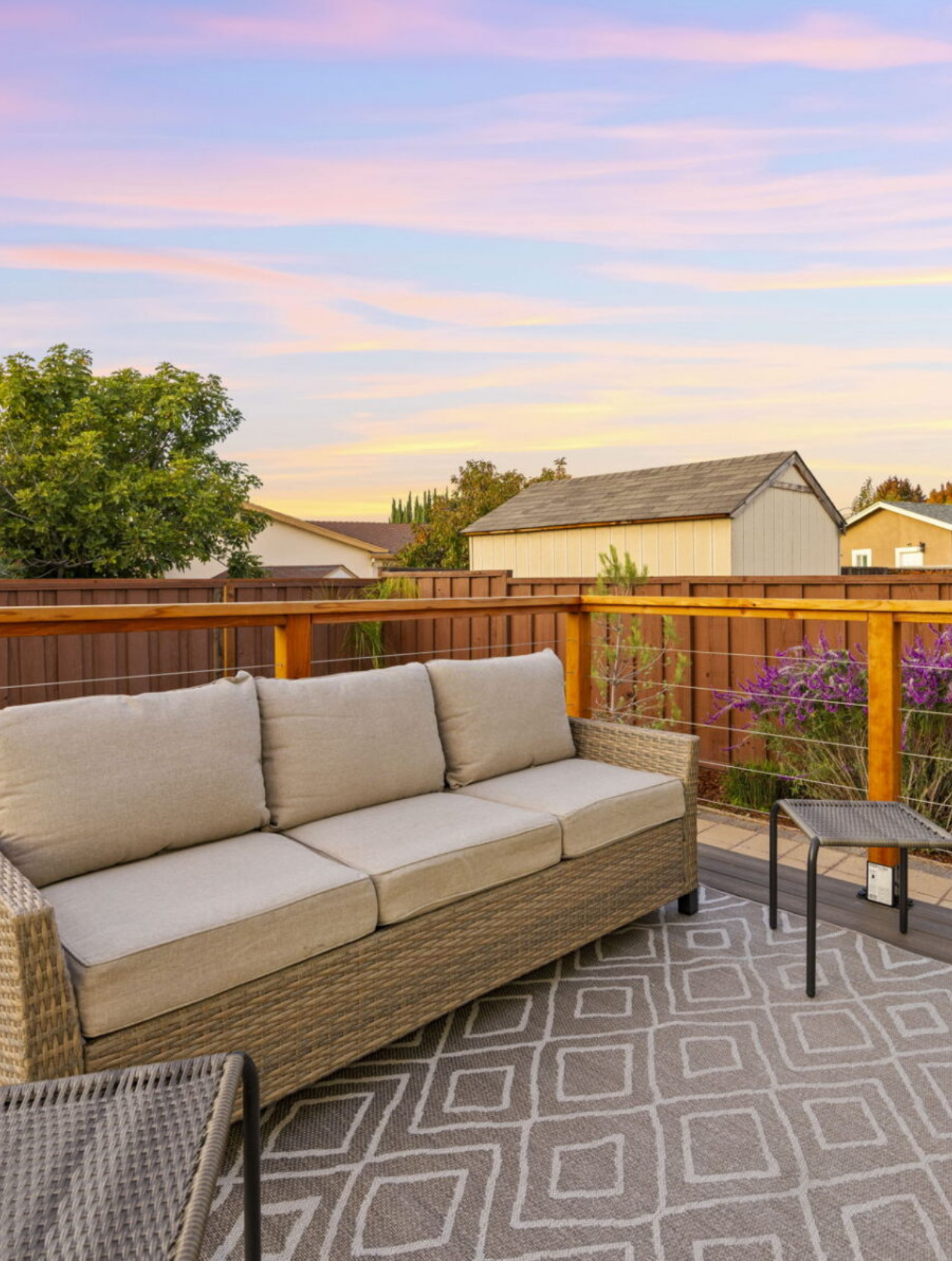 Outdoor patio with a wicker sofa, cushioned seating, two side tables, patterned rug, wooden fence, and plants, with a sunset sky in the background.