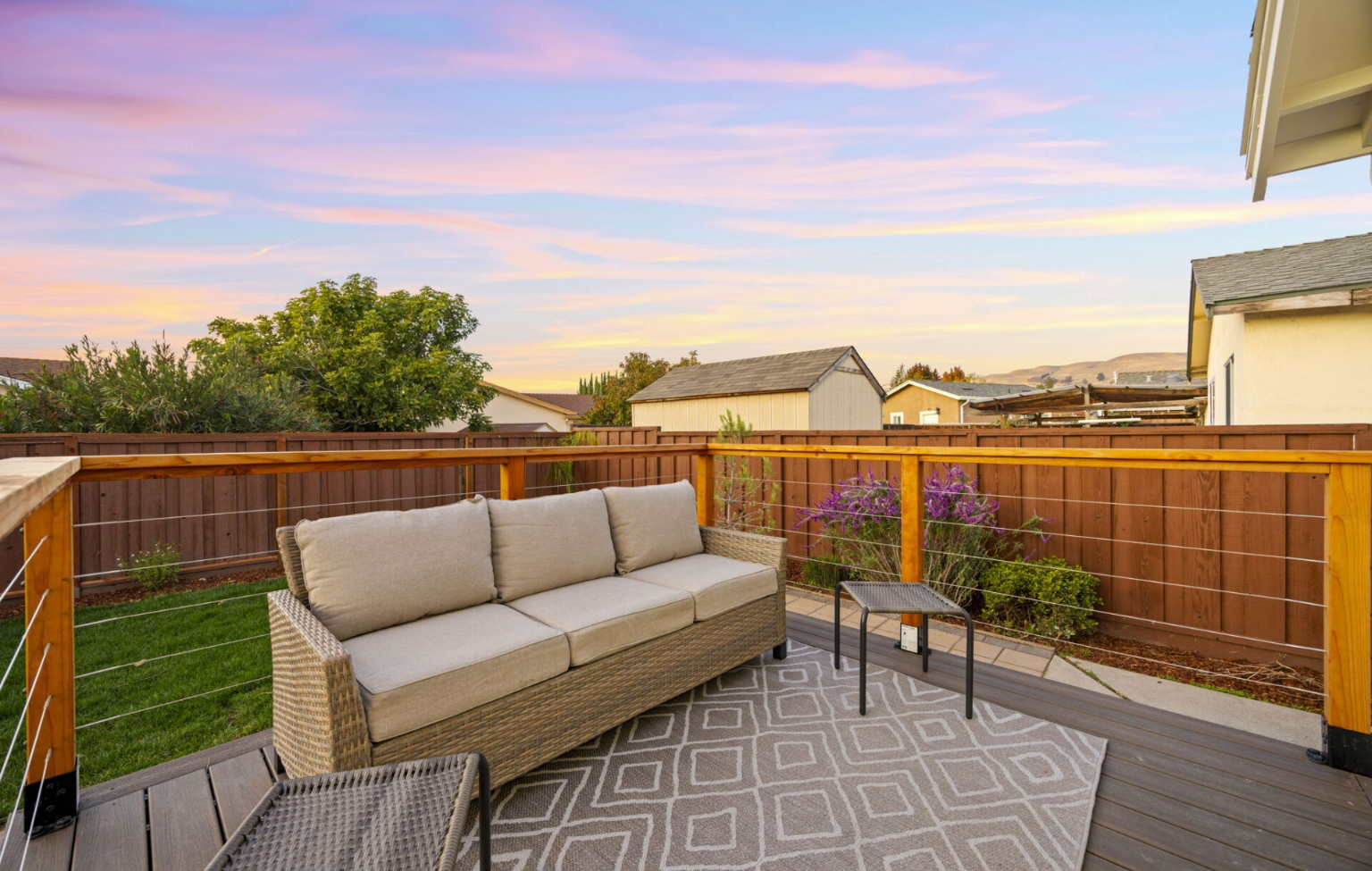 Outdoor patio with wicker sofa, small table, and chair on a patterned rug, surrounded by a wooden fence and garden, under a colorful evening sky.