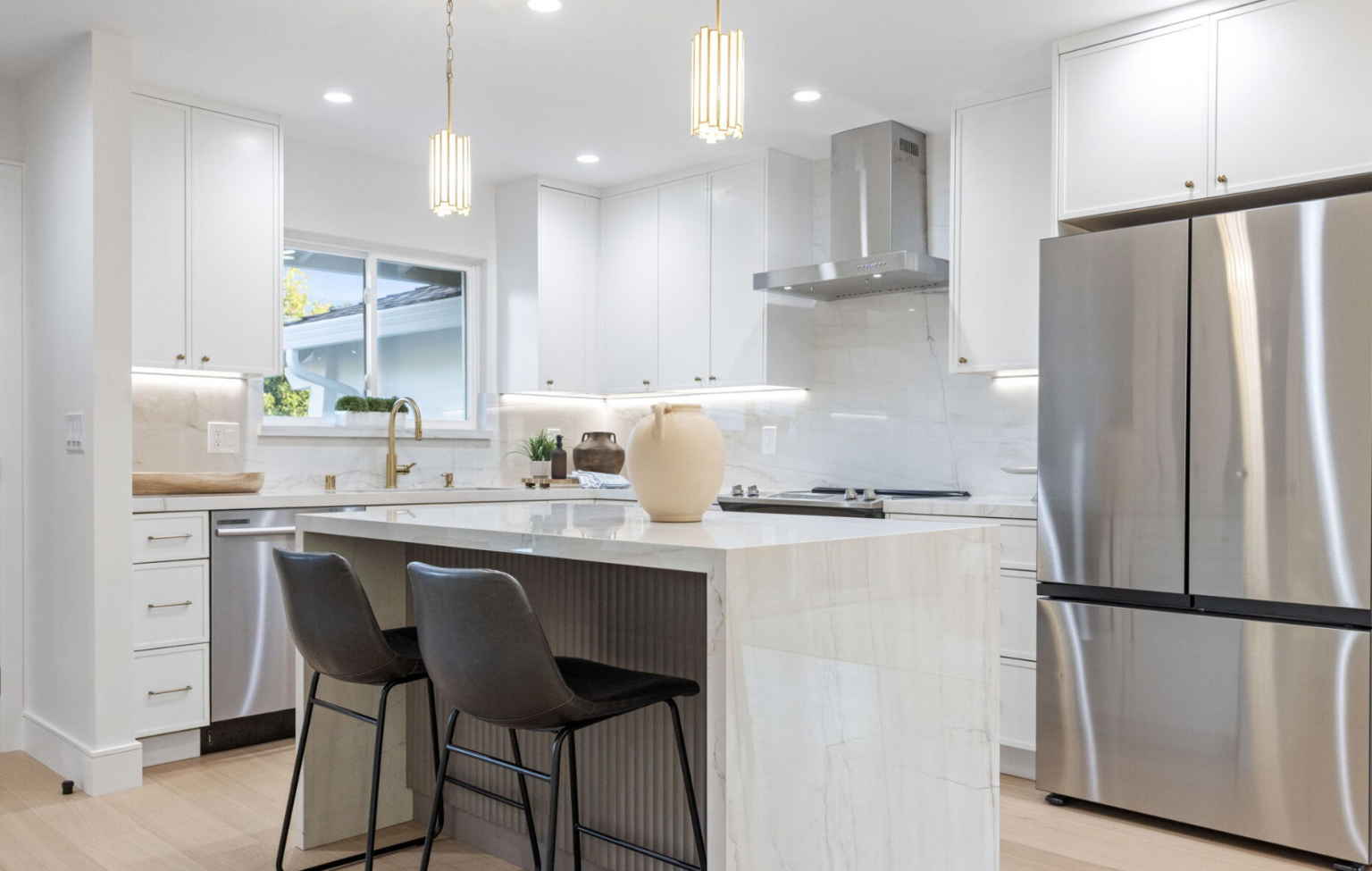 Modern kitchen with white cabinetry, stainless steel appliances, marble island with two black chairs, and decorative vase centerpiece.