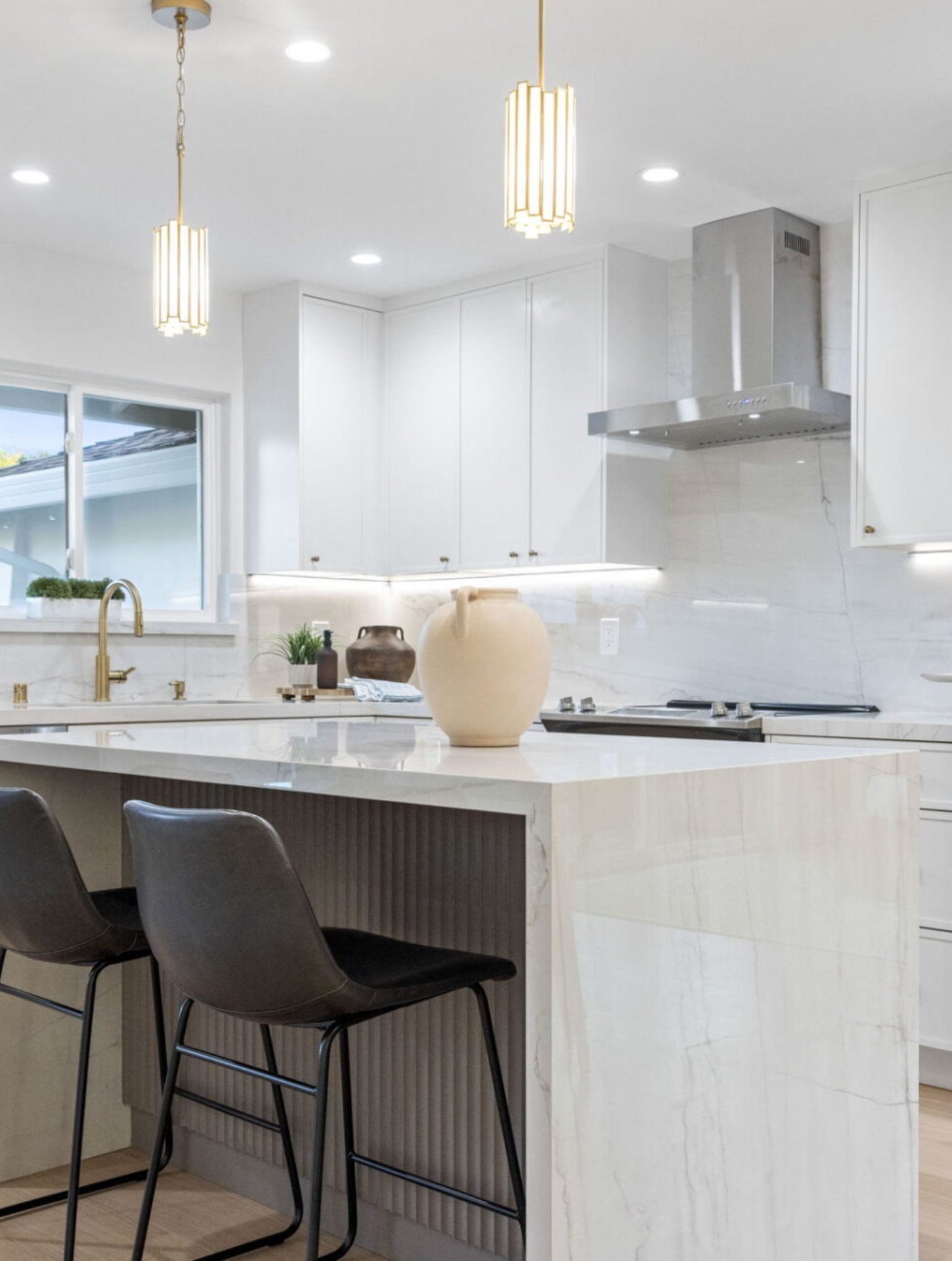 Modern kitchen with white marble island, two black chairs, gold fixtures, pendant lights, a range hood, and minimal decor including a beige vase on the island.