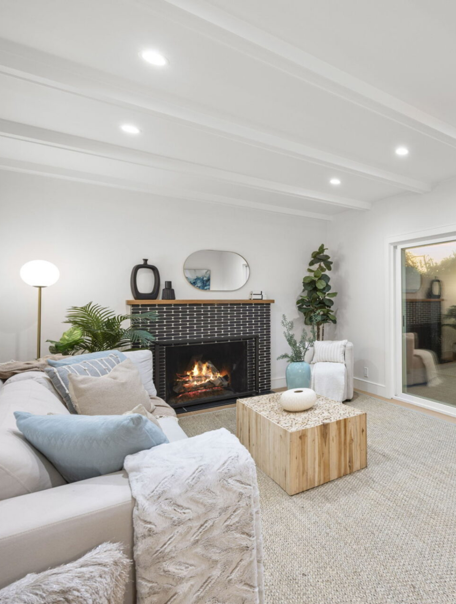 A modern living room with a lit fireplace, neutral-toned sofa, light wood coffee table, indoor plants, and large glass doors letting in natural light.
