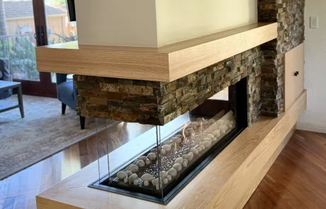 A modern double-sided glass fireplace with stone and wood accents, surrounded by white stones, separates the living area from an adjacent room.