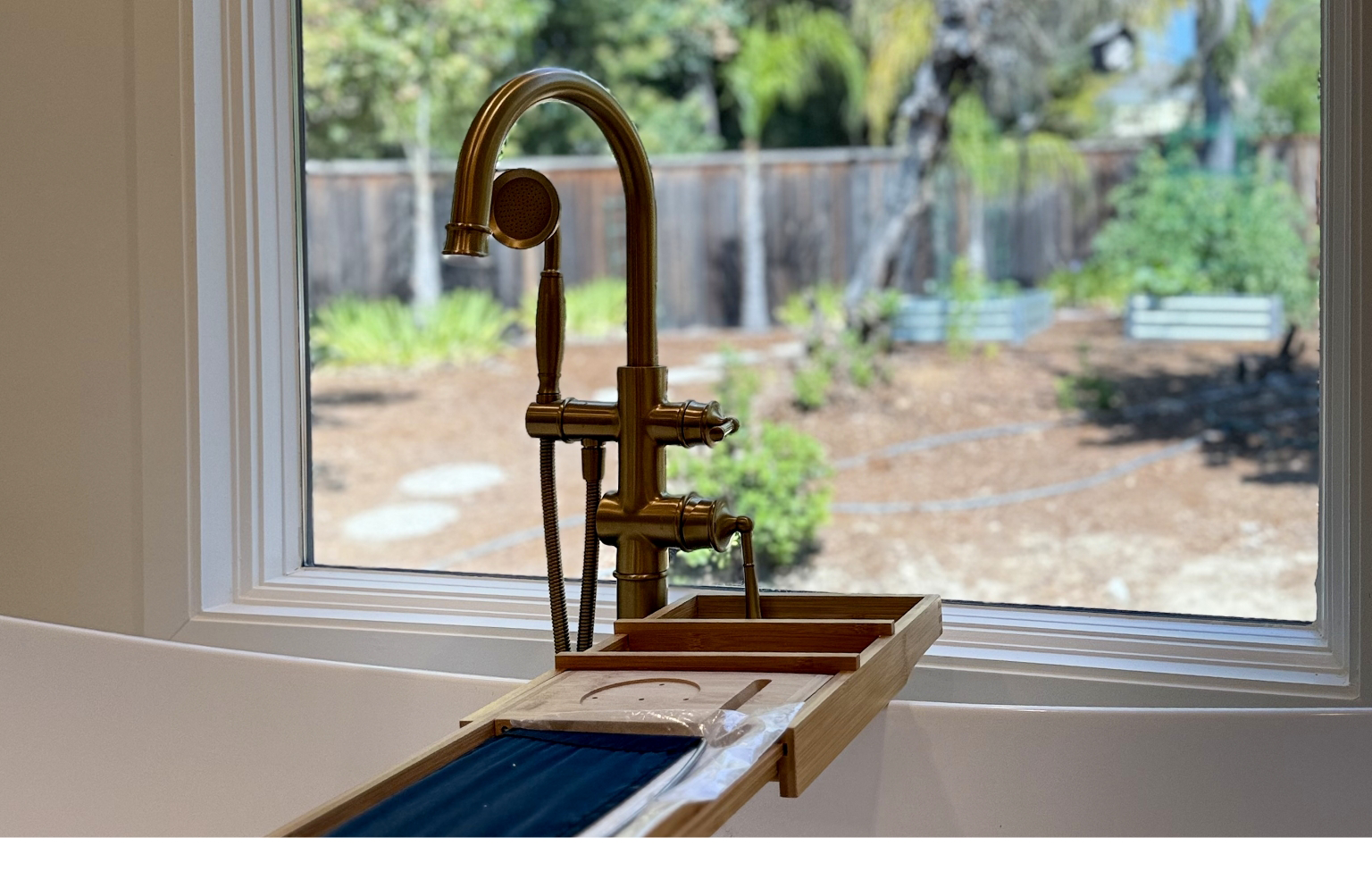 A modern brass bathtub faucet is mounted by a window, with a wooden bath caddy resting on the tub and a garden visible outside—a serene touch in this Full Interior Remodel With Custom Kitchen in Danville.
