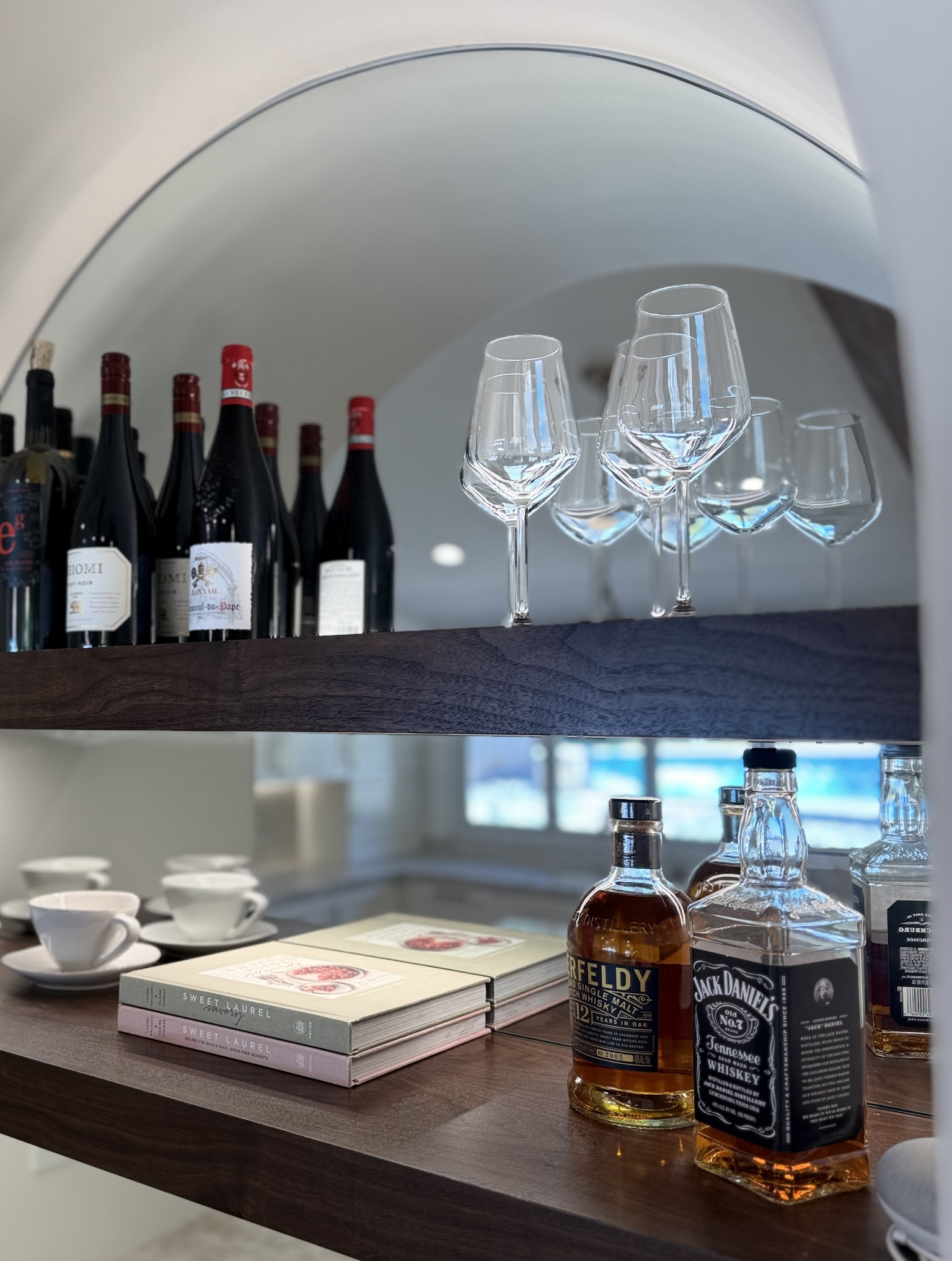 A shelf with wine bottles, empty wine glasses, whiskey bottles, teacups, and books arranged neatly in a well-lit room, showcasing the elegance of a full interior remodel with custom kitchen in Danville.
