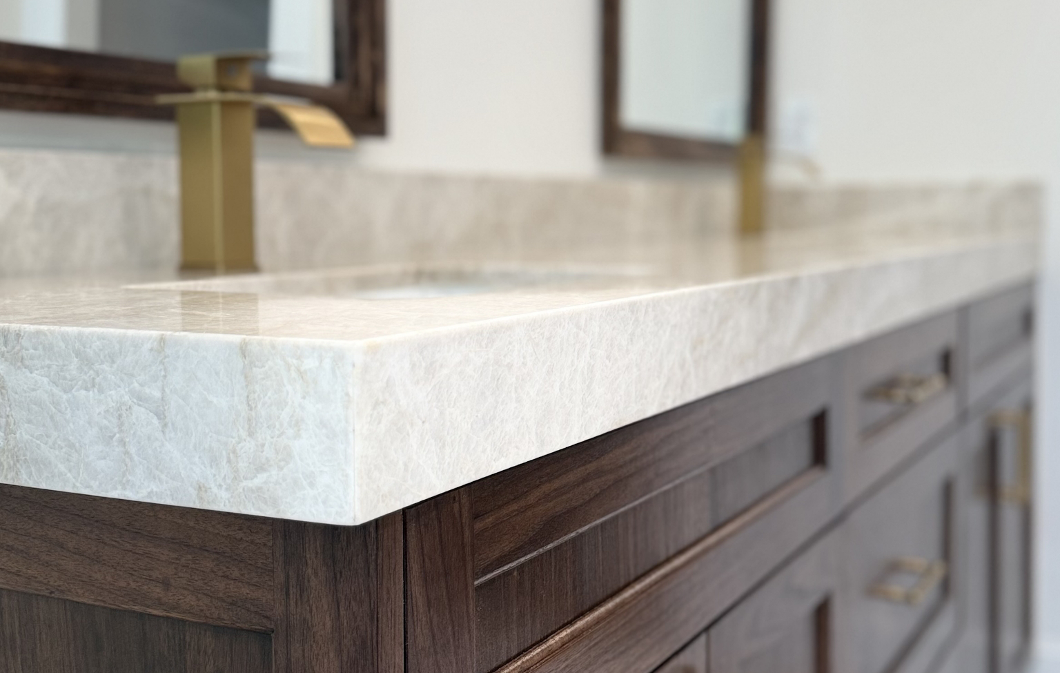Close-up of a bathroom vanity with a light marble countertop, wooden drawers, and brass fixtures, part of a full interior remodel with custom kitchen in Danville; a blurred mirror and faucet appear in the background.