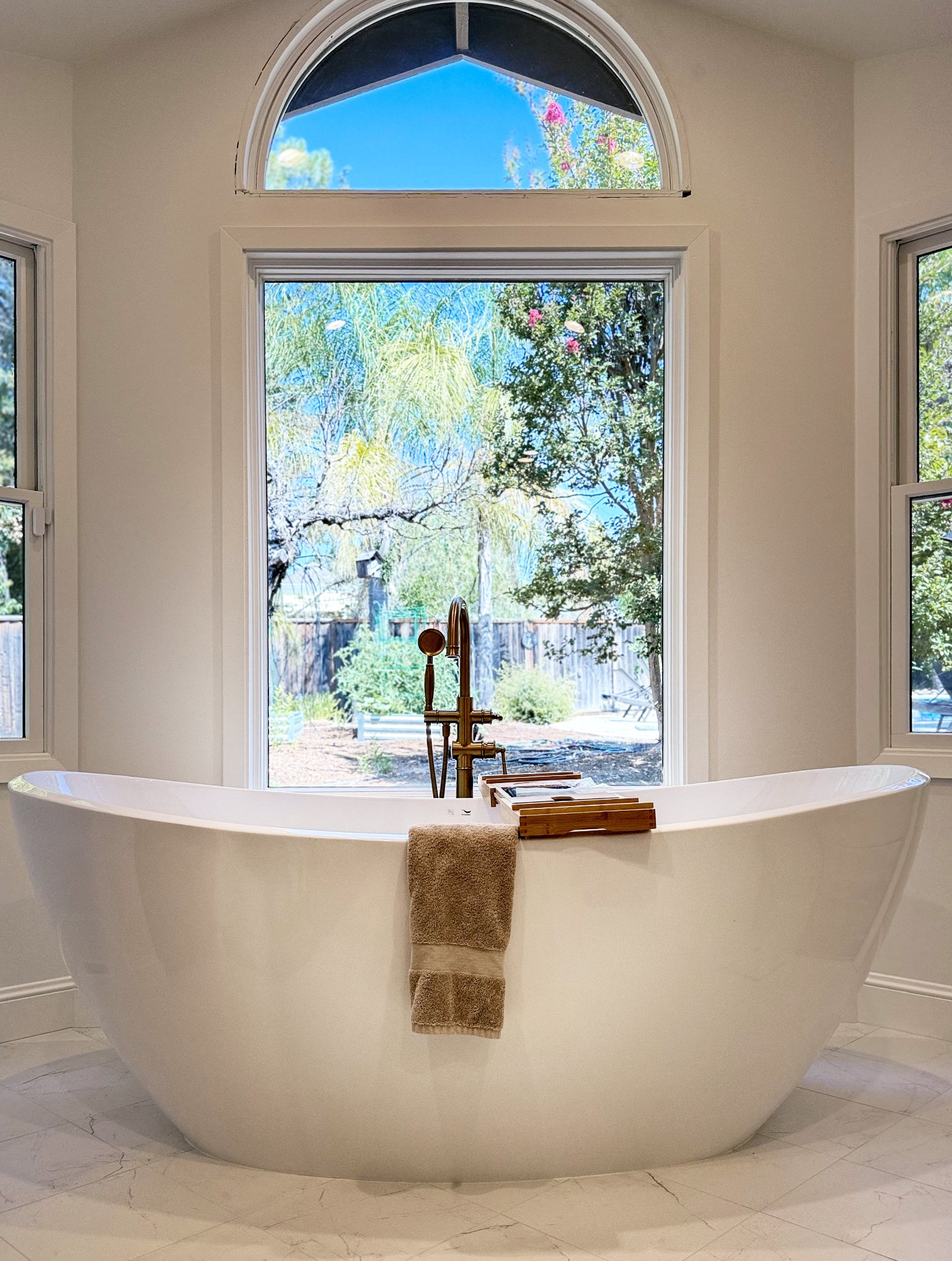 A white freestanding bathtub sits under a large window with a view of a green backyard; part of a full interior remodel with custom kitchen in Danville, a brown towel hangs on the tub and a tray rests on the edge.