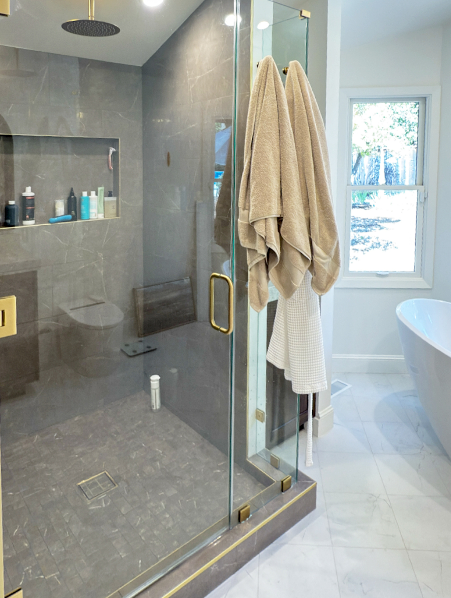 Modern bathroom with a glass-enclosed shower, gray tile walls, shelves with toiletries, and a white freestanding bathtub near a window—part of a full interior remodel with custom kitchen in Danville.