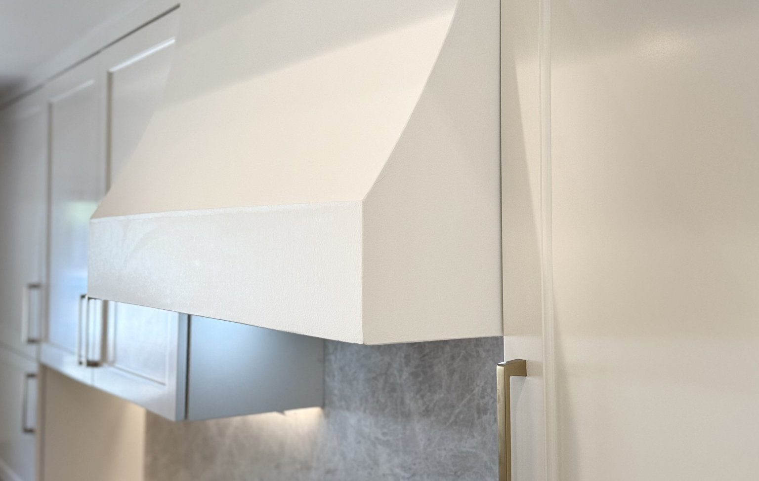 Close-up of a white kitchen range hood and adjacent cabinets with gold handles against a light-colored backsplash from a Full Interior Remodel With Custom Kitchen in Danville.