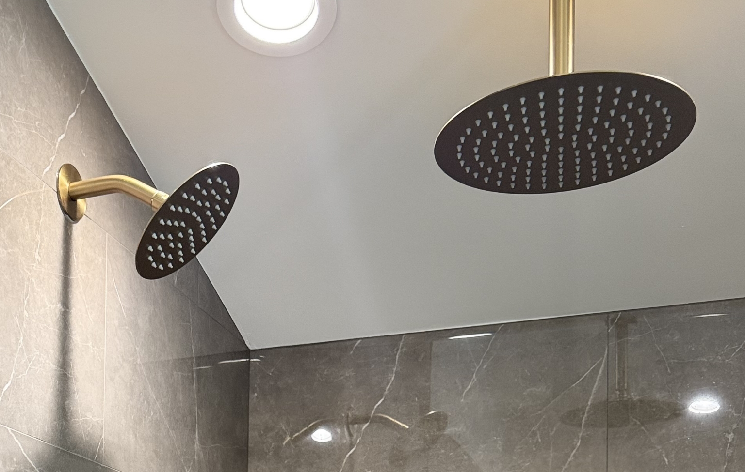 Two black round showerheads with gold arms are mounted on a gray tiled wall and white ceiling, under a recessed ceiling light—a stunning feature from a Full Interior Remodel With Custom Kitchen in Danville.