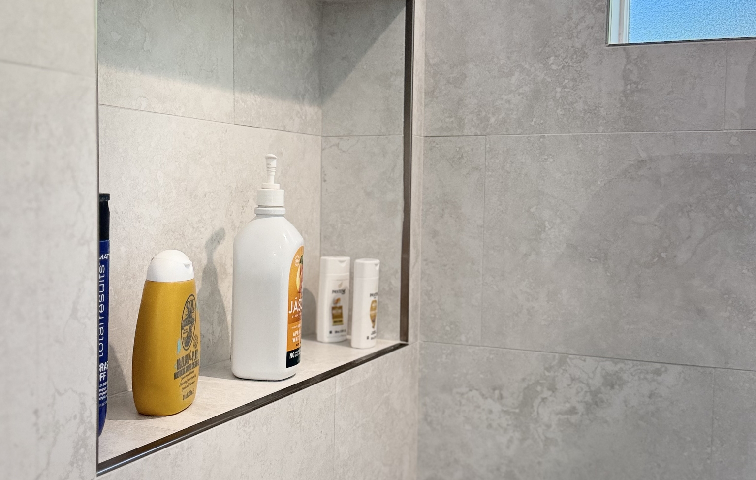 Shower shelf with various bath products, including bottles of shampoo, conditioner, lotion, and toothpaste, in a stylish space—part of a Full Interior Remodel With Custom Kitchen in Danville—set against a light gray tiled wall.