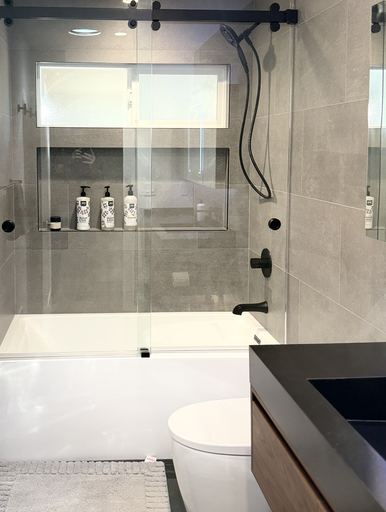 Modern bathroom with a glass-enclosed shower, black fixtures, a white bathtub, and a sleek dark vanity—part of a Full Interior Remodel With Custom Kitchen in Danville. Wall-mounted shelf holds toiletries beside the stylish white toilet.