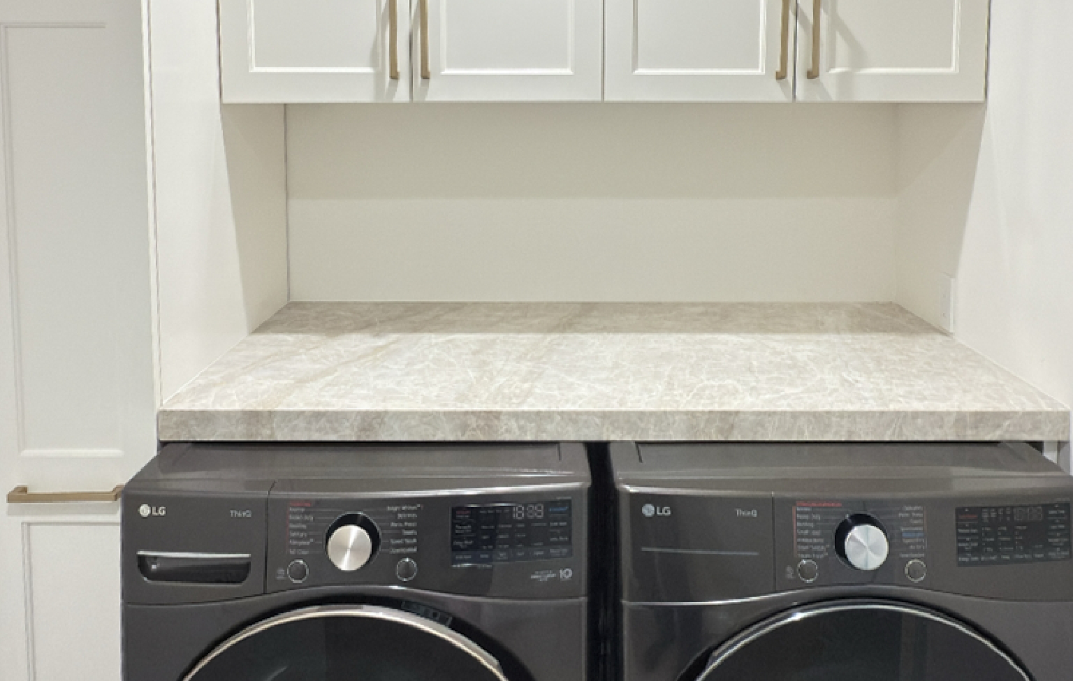 Two front-loading LG washers and dryers sit beneath a stone-look countertop, with white cabinets and gold handles mounted above—part of a full interior remodel with custom kitchen in Danville.