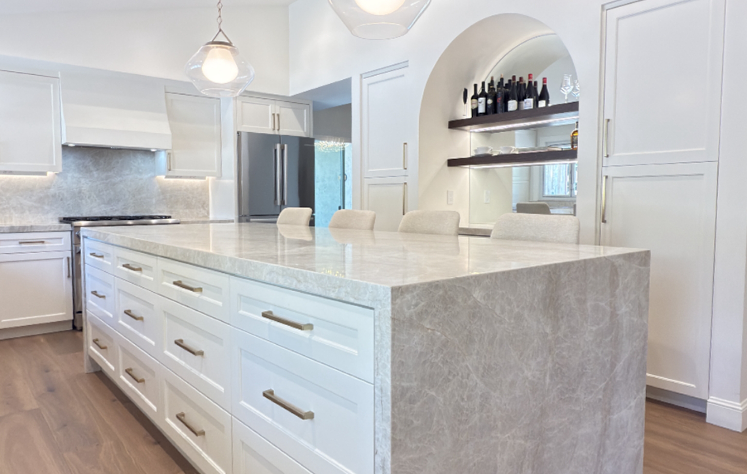 Modern kitchen from a full interior remodel with custom kitchen in Danville features a large marble island, white cabinets, stainless steel appliances, pendant lights, and shelves with wine bottles and glasses in an arched nook.