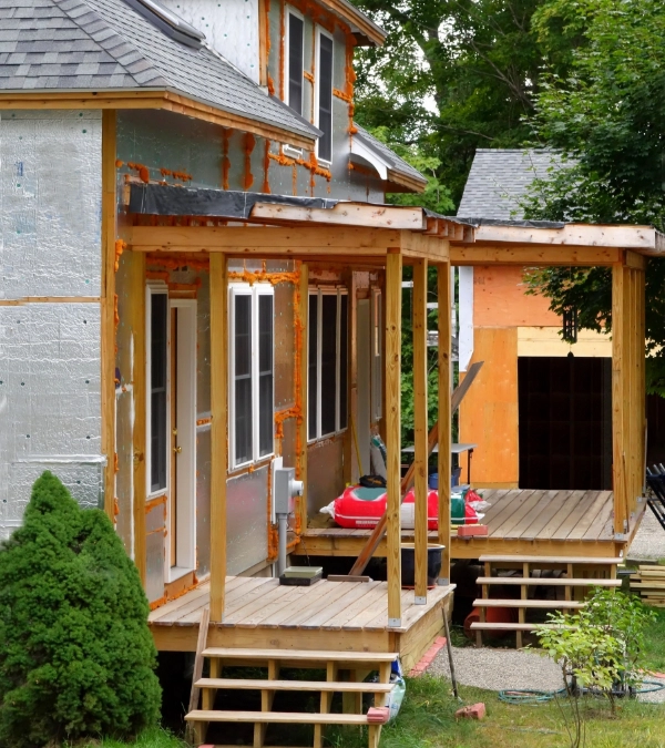 Wooden porch under construction on a partially renovated house, with exposed insulation and construction materials visible.