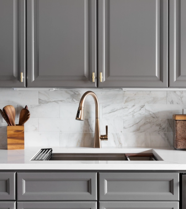Modern kitchen with grey cabinets, marble backsplash, white countertop, bronze faucet, wooden utensil holder, and a brown storage container.
