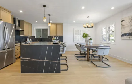 Modern kitchen and dining area with light wood floors, a black marble island, stainless steel appliances, beige cabinets, gray chairs, and contemporary lighting—perfect inspiration for Bay Area Remodeling Projects.