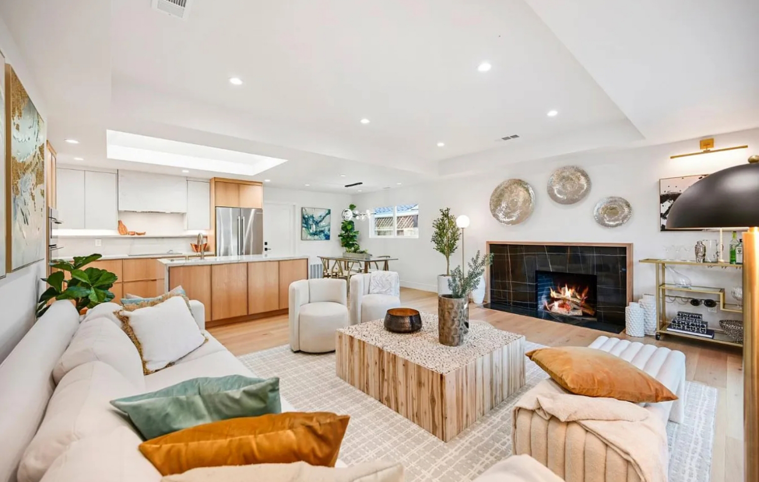A modern living room with a white sectional sofa, wooden coffee table, fireplace, wall art, and an open view to a kitchen with wood cabinetry. The space is bright with natural and recessed lighting, showcasing Bay Area Remodeling Projects.