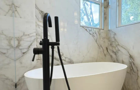 Modern bathroom with a white freestanding tub, black floor-mounted faucet, and marble walls. Two windows above let in natural light, showcasing Bay Area Remodeling Projects.