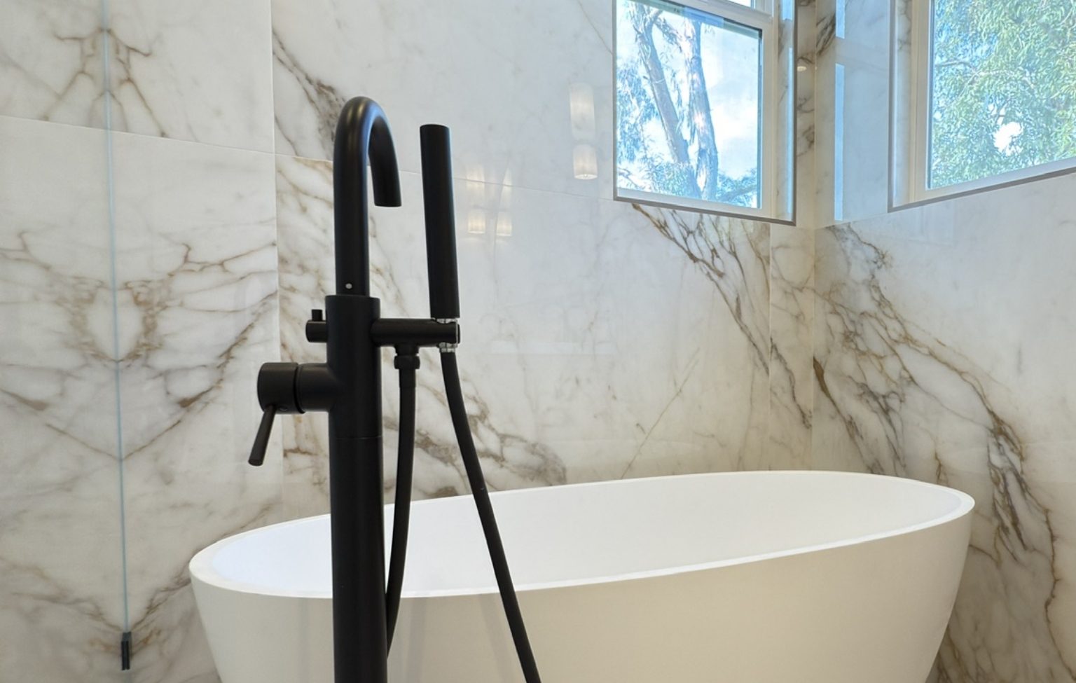 Modern bathroom with a white freestanding tub, black floor-mounted faucet, and marble walls. Two windows above let in natural light, showcasing Bay Area Remodeling Projects.