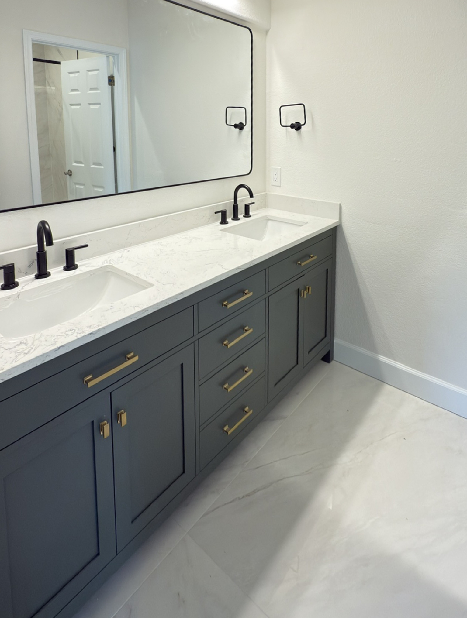 Bathroom with a large mirror, double sinks, black faucets, gray vanity cabinets with gold handles, and white marble countertop on a tiled floor.