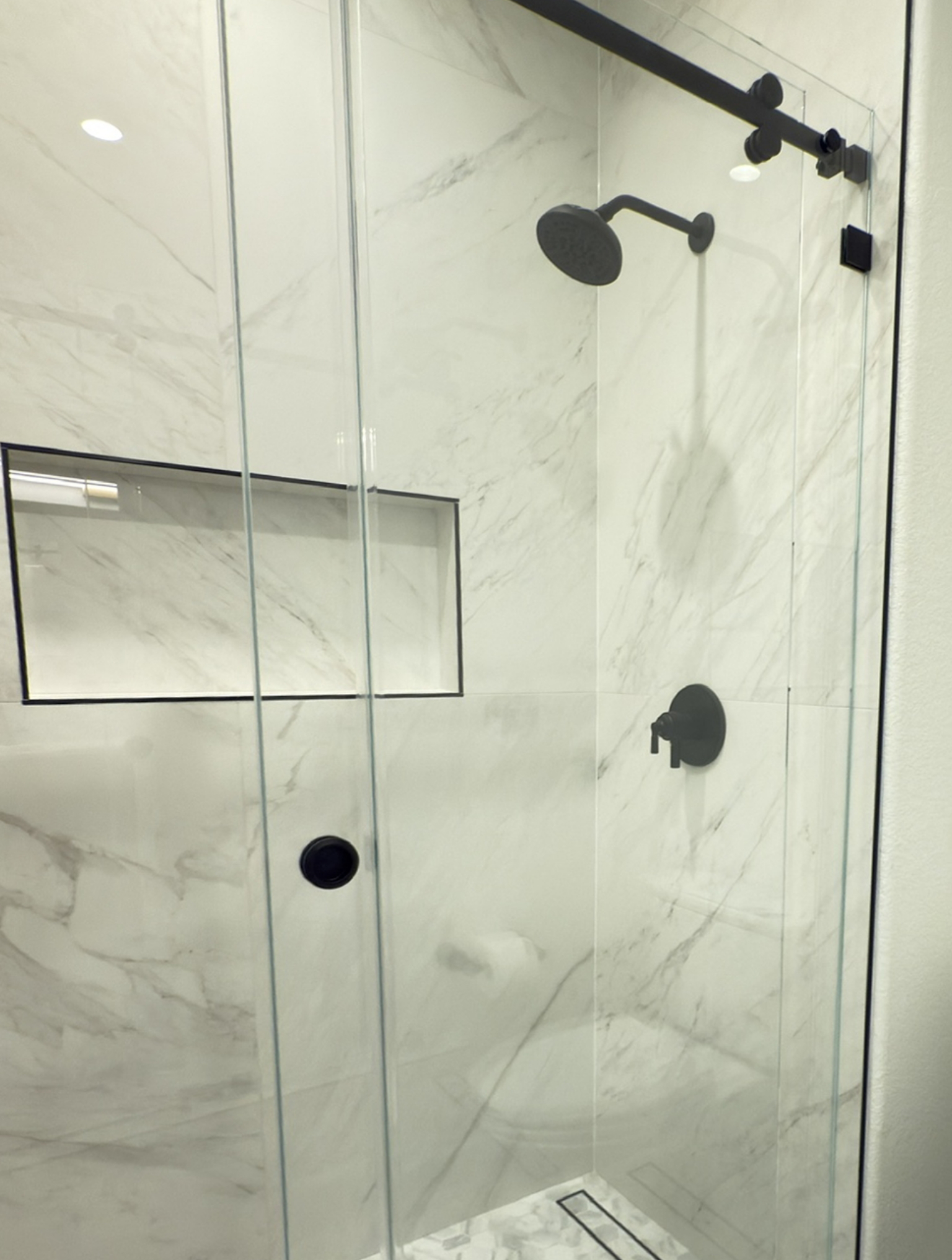 Modern shower with glass doors, marble-patterned walls, a built-in shelf, and matte black fixtures including a rainfall showerhead and handle.