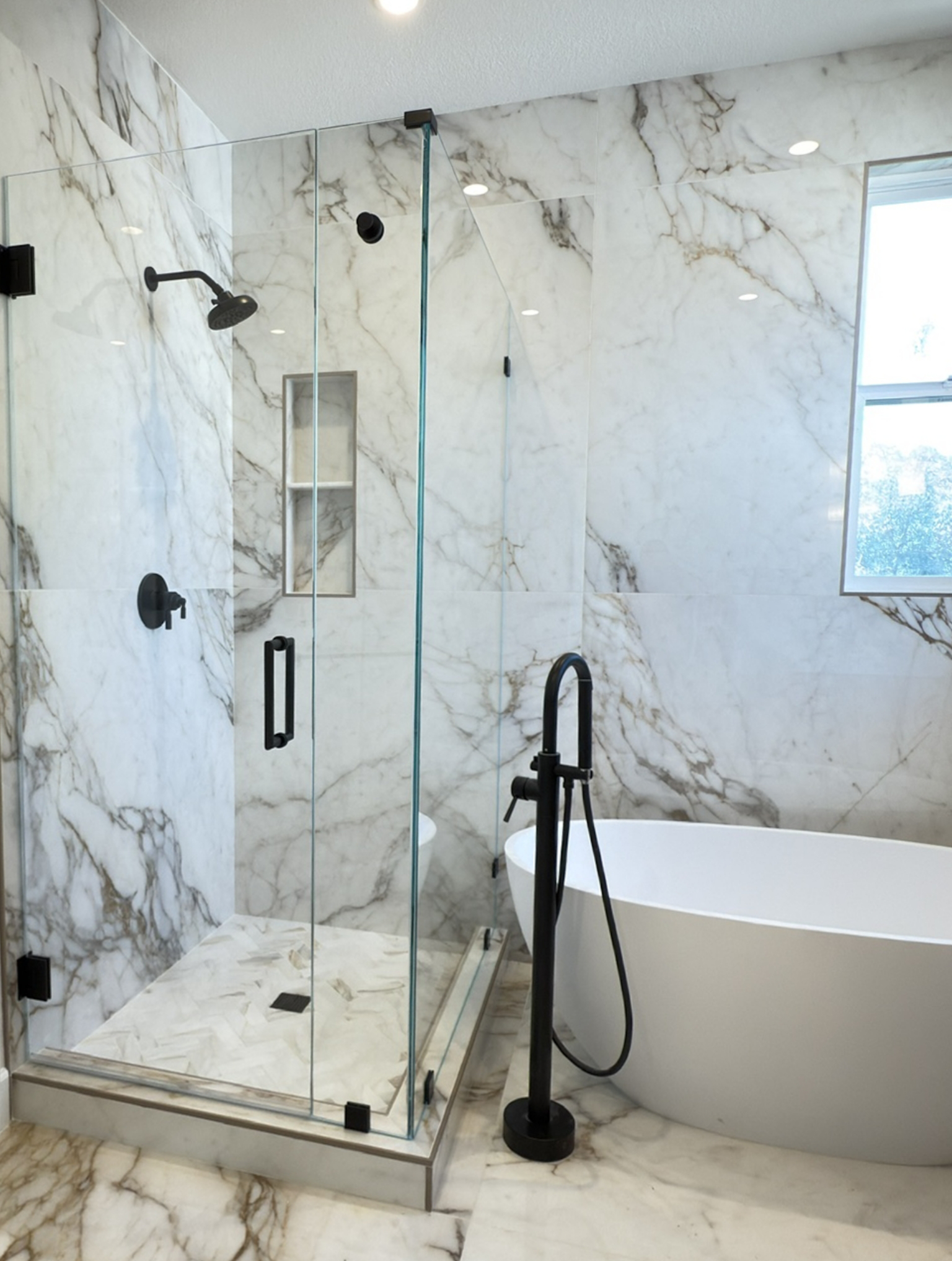 Modern bathroom with marble walls and floors, featuring a glass-enclosed shower with black fixtures and a freestanding white bathtub next to a window.
