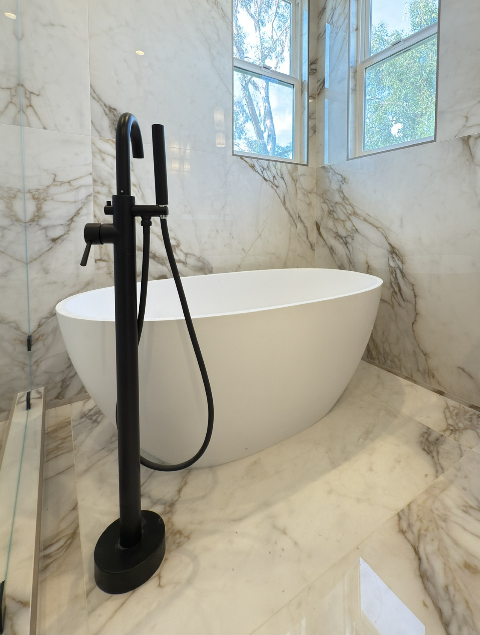 Modern bathroom with a freestanding white oval bathtub, black floor-mounted faucet, and marble walls and floor. Two windows provide natural light.