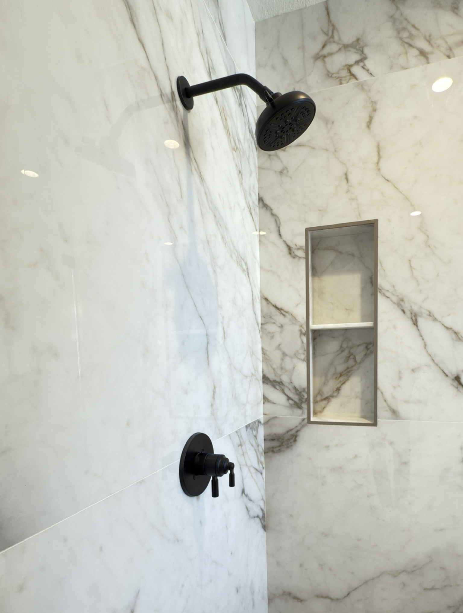 A black showerhead and handle are mounted on a marble-patterned wall with a built-in recessed shelf visible in the background.