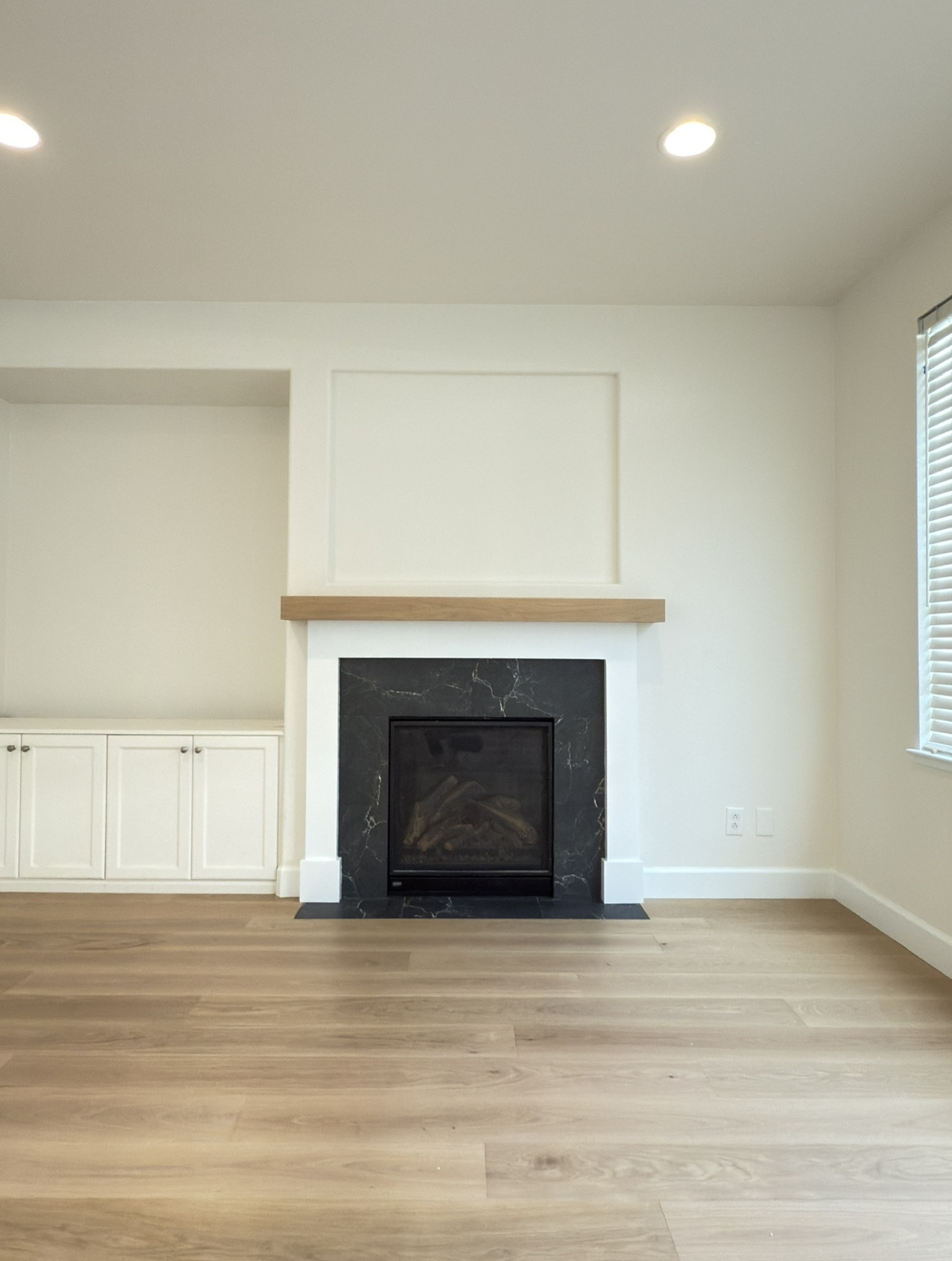 A modern living room with light wood flooring, a black fireplace with a wood mantel, built-in white cabinets, and a window with blinds.
