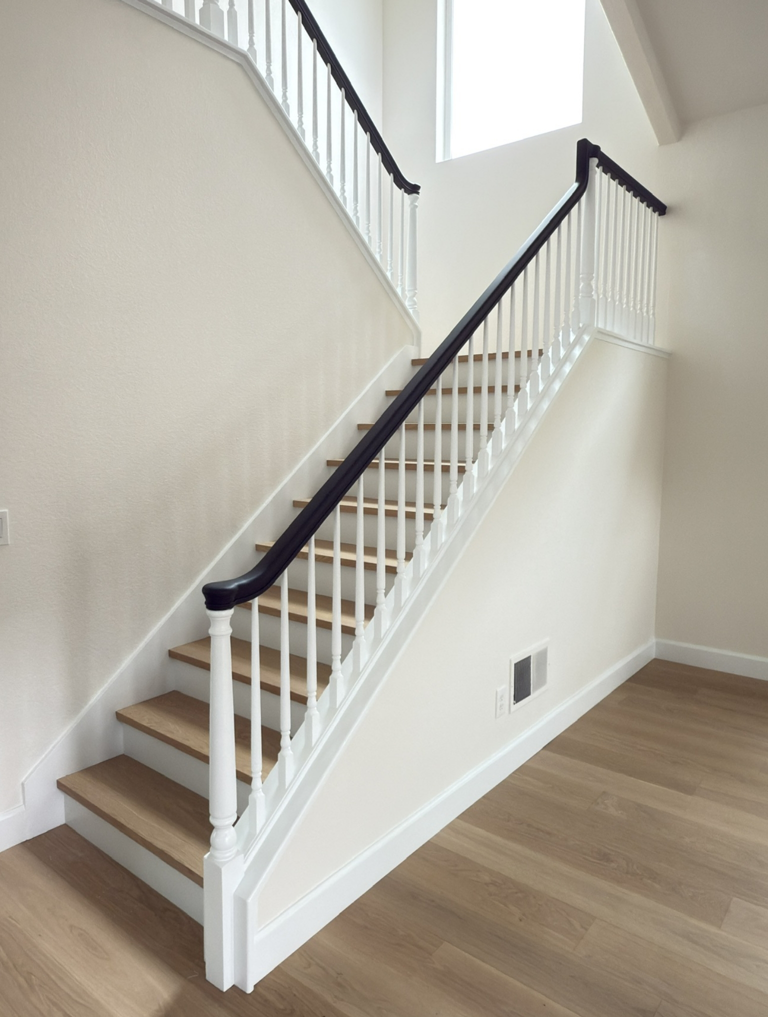A wooden staircase with white railings and a dark handrail, set against a light-colored wall in a room with light wood flooring.