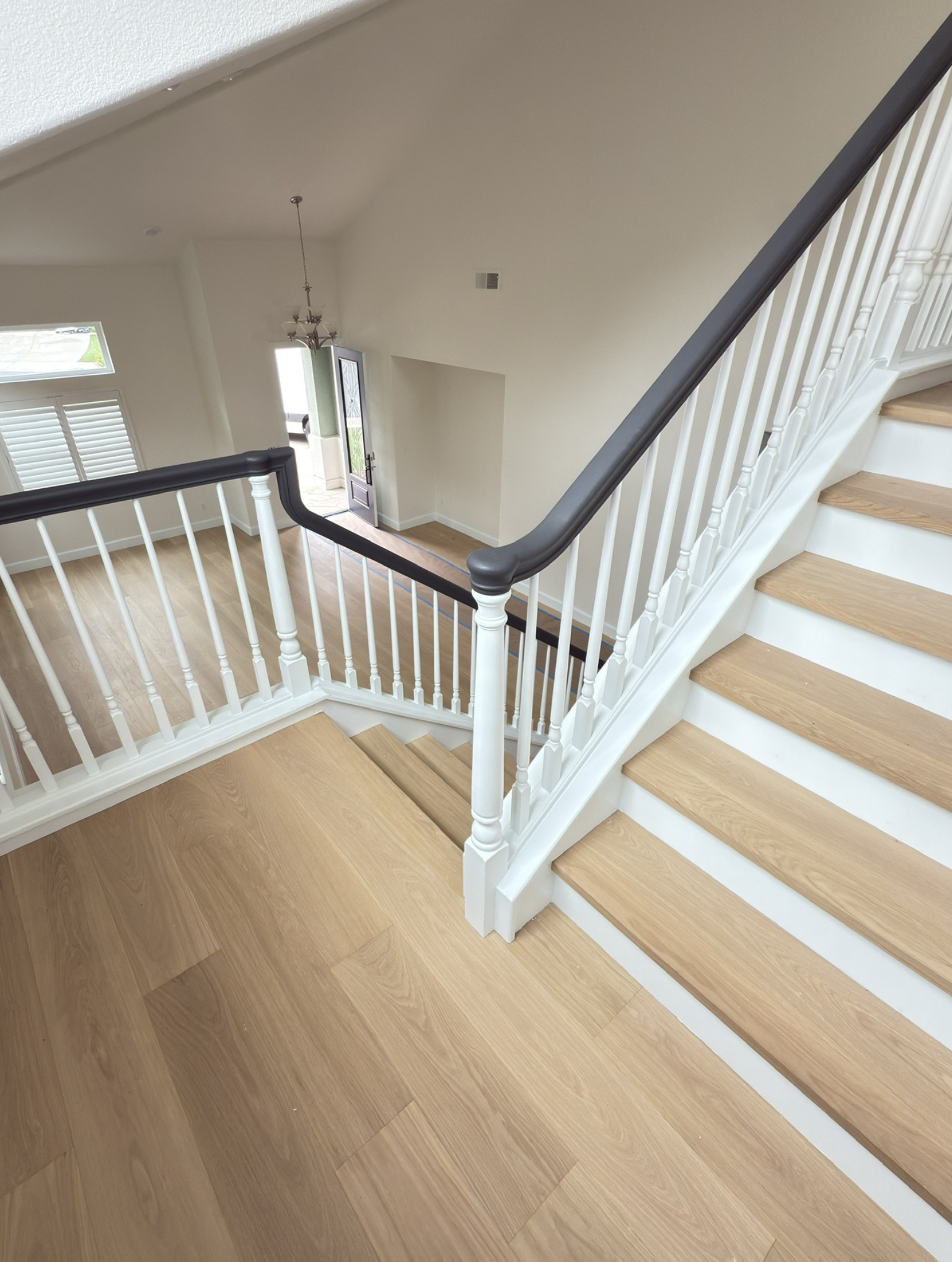 A view from the top of a staircase with light wood steps and white railing, overlooking an open foyer with large windows and a chandelier.