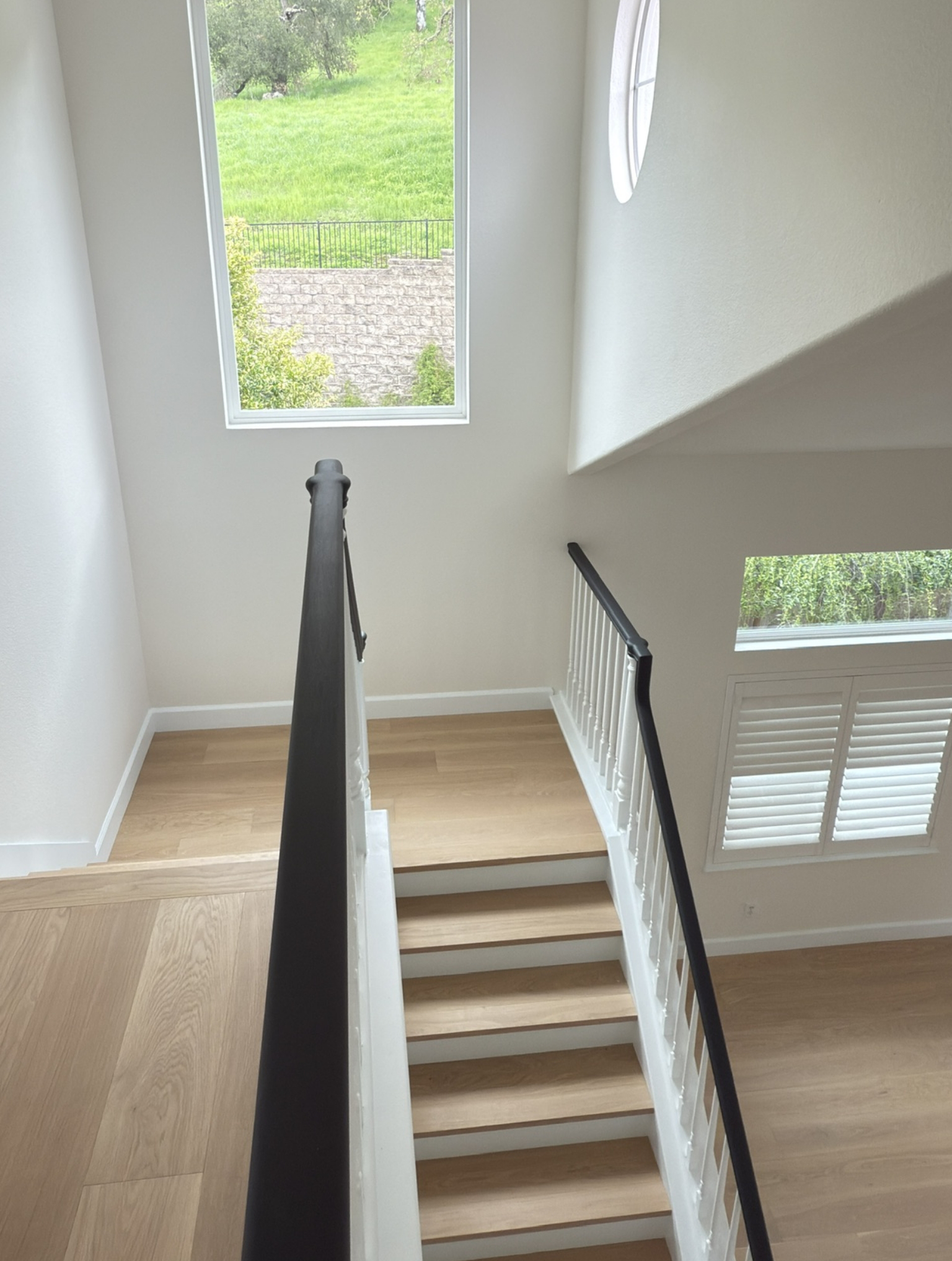 Interior view of a staircase with white walls, wooden steps, black handrails, large rectangular window, and round window, overlooking a green outdoor area.